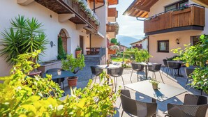An outdoor patio with tables and chairs, surrounded by potted plants and wooden balconies. - Rebenheim Frühstückspension - FEWO (Termeno Sulla Strada del Vino)