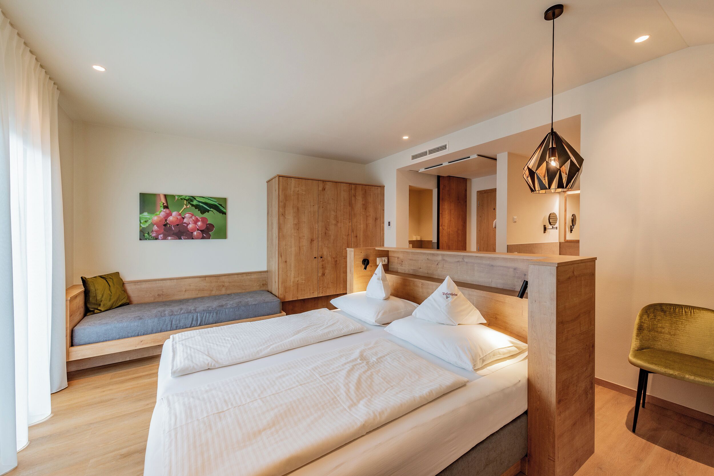 A modern bedroom with a large bed, a wooden headboard, a bench with cushions, and a hanging light fixture.