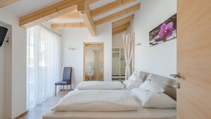 A bedroom with a wooden ceiling, a bed with white bedding, a chair, and a bathroom visible through an open door. - Rebenheim Frühstückspension - FEWO (Termeno Sulla Strada del Vino)