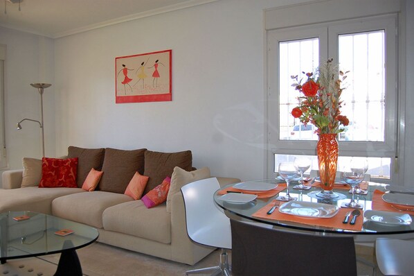 A living room with a sofa, a glass coffee table, a dining table with chairs, a vase with flowers, and a painting on the wall.