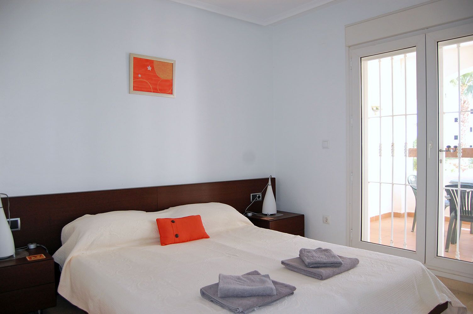 A neatly made bed with a white bedspread, a red pillow, and two folded towels. There is a nightstand with a lamp and a small table beside the bed. A framed picture hangs on the wall above the bed.