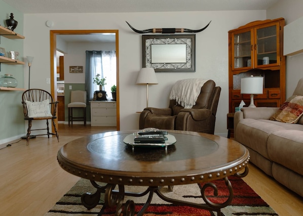 A living room with a round wooden coffee table, a brown leather armchair, a beige sofa, and a wooden cabinet with glass doors.