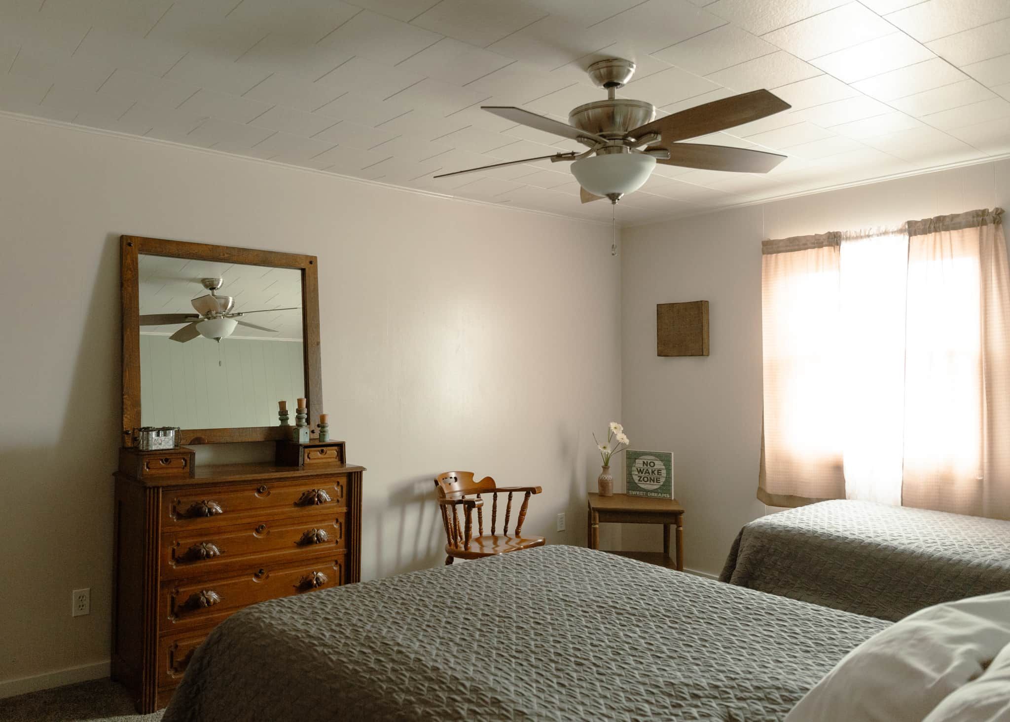 Ein Schlafzimmer mit zwei Betten, einem Schminktisch, einem Stuhl, einem Deckenventilator und einem Fenster mit Vorhängen.