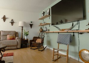 Una sala de estar con un sofá, un sillón mecededor, una mesa y un televisor montado en la pared.