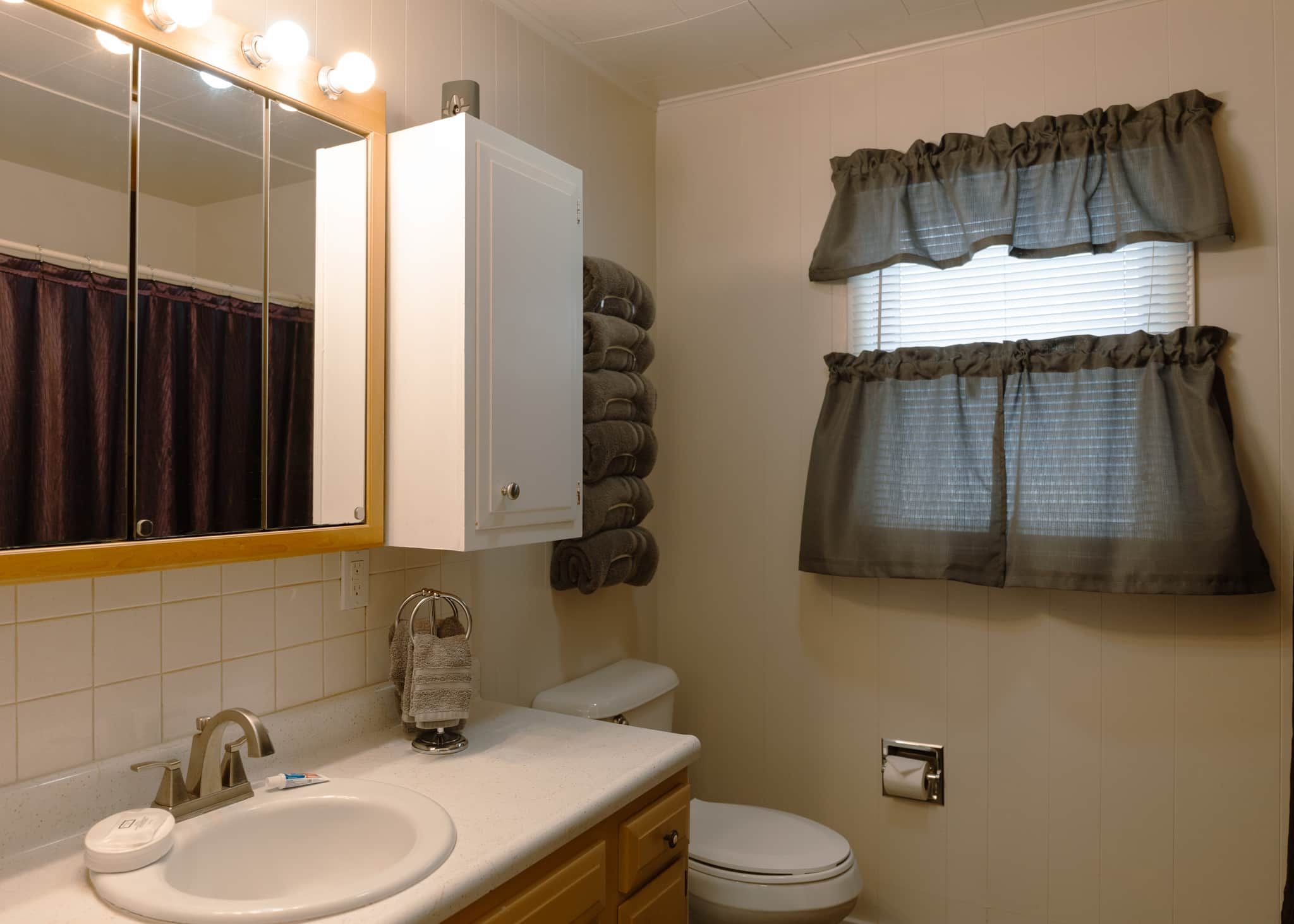 Un baño con lavabo, espejo, inodoro, toallero y ventana con cortinas.