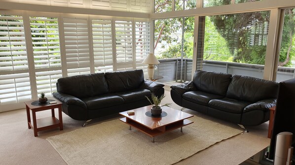 A living room with two black leather sofas, a wooden coffee table with a plant, and a lamp. Large windows with blinds are visible.