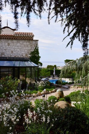 A stone building with a glass conservatory, a pool, and a garden with white flowers and rocks.
