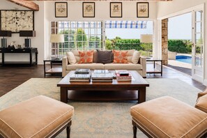 A spacious living room with a sofa, coffee table, and ottomans, a dining area with a table and chairs, and a view of a swimming pool.