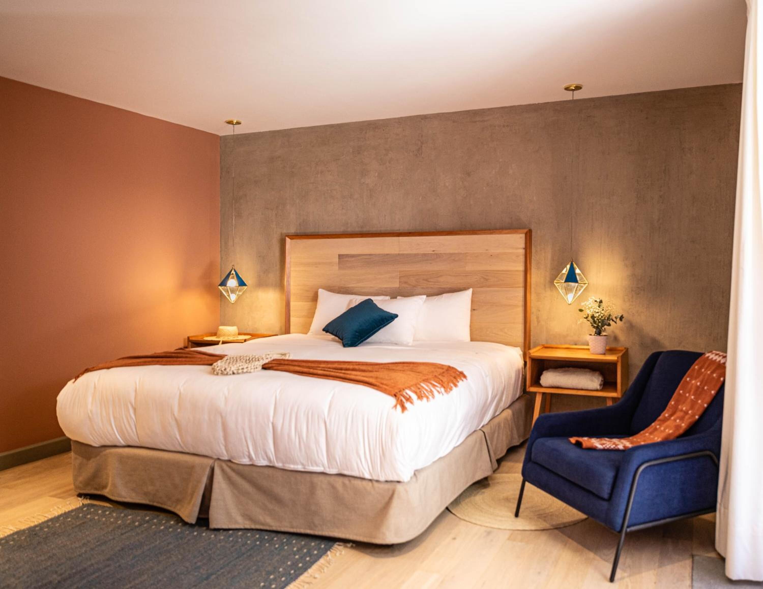 Una habitación de hotel moderna con una cama grande, un sillón azul y una mesita de noche de madera con una lámpara.