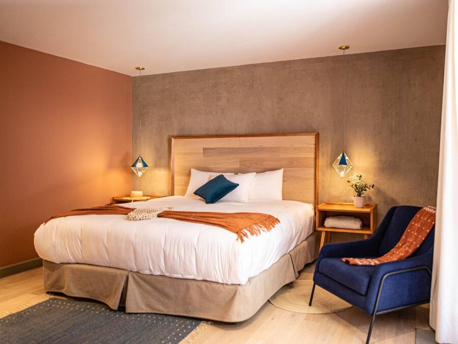 Una habitación de hotel moderna con una cama grande, un sillón azul y una mesita de noche de madera con una lámpara.