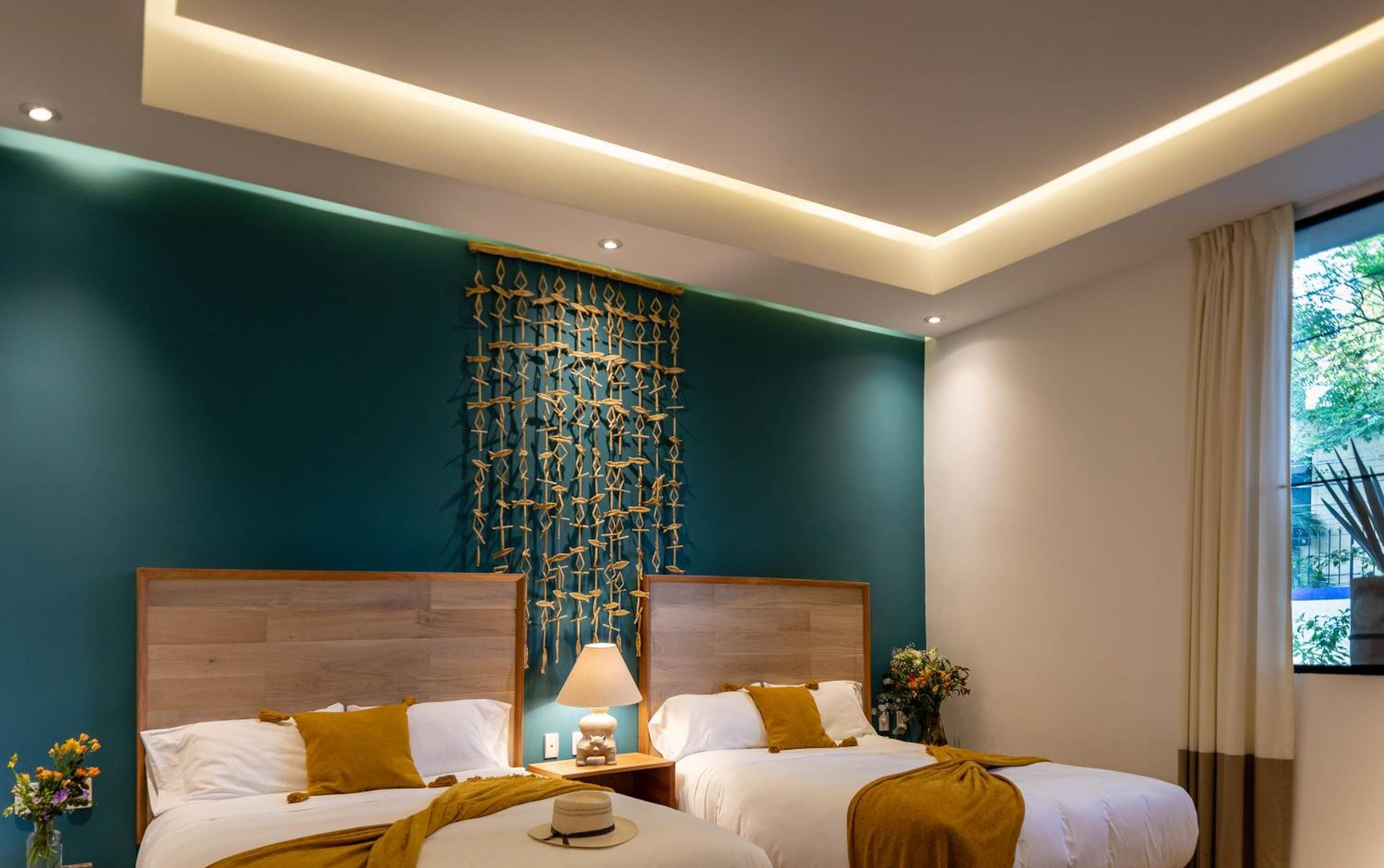Una habitación de hotel moderna con dos camas, un tapiz decorativo, una mesita de noche con lámpara y una ventana con cortinas.