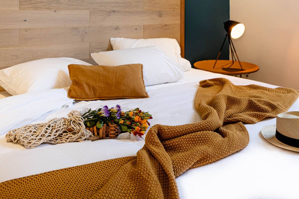 A neatly made bed with white linens, a brown knitted blanket, a woven throw, a straw hat, and a small table with a lamp beside it.