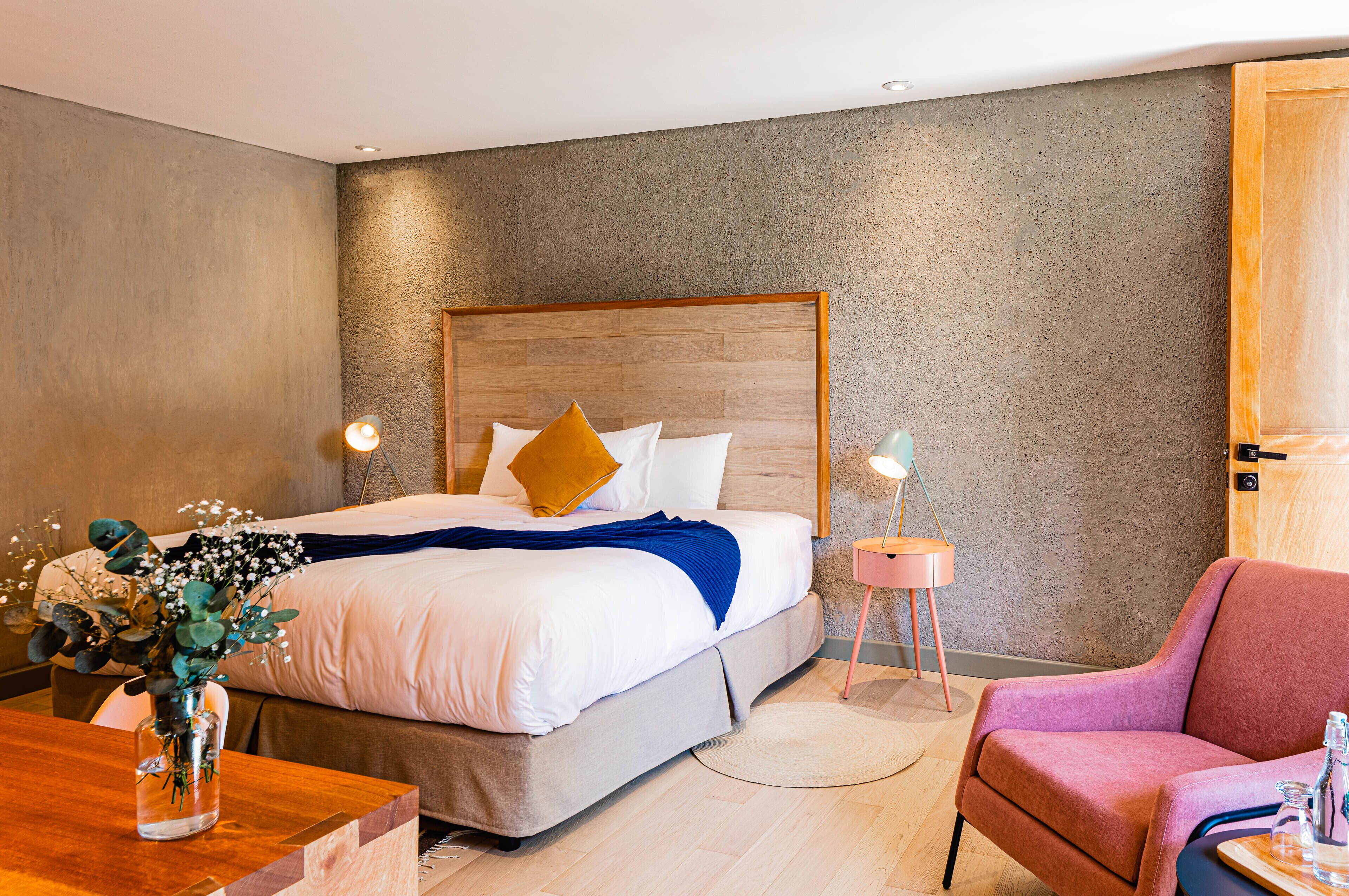 A modern hotel room with a large bed, a wooden headboard, a pink armchair, and a small side table with a lamp.
