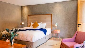A modern hotel room with a large bed, a wooden headboard, a pink armchair, and a small side table with a lamp.