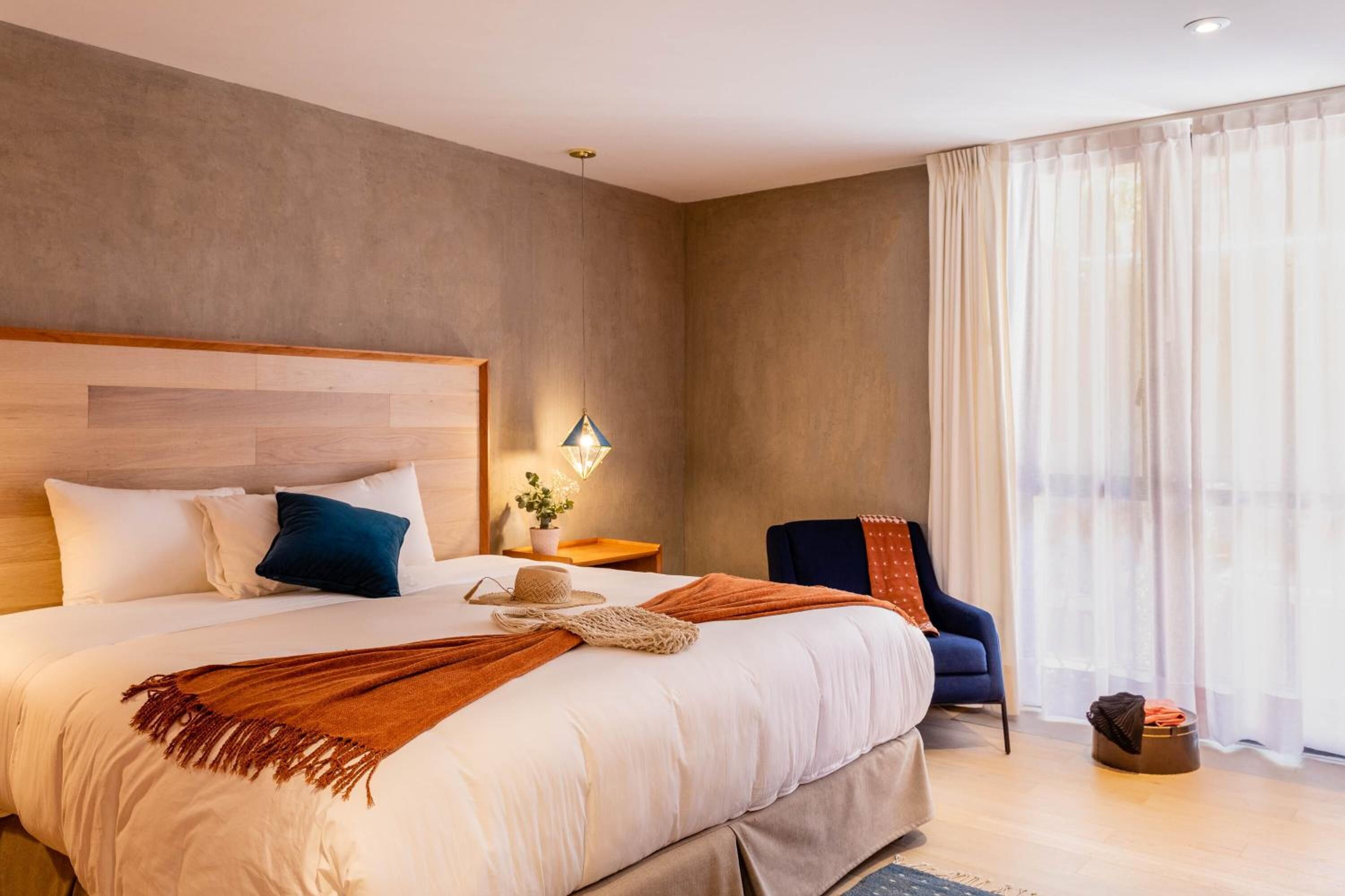 Une chambre d’hôtel moderne avec un grand lit, une table de chevet, une chaise et une fenêtre avec des rideaux légers.
