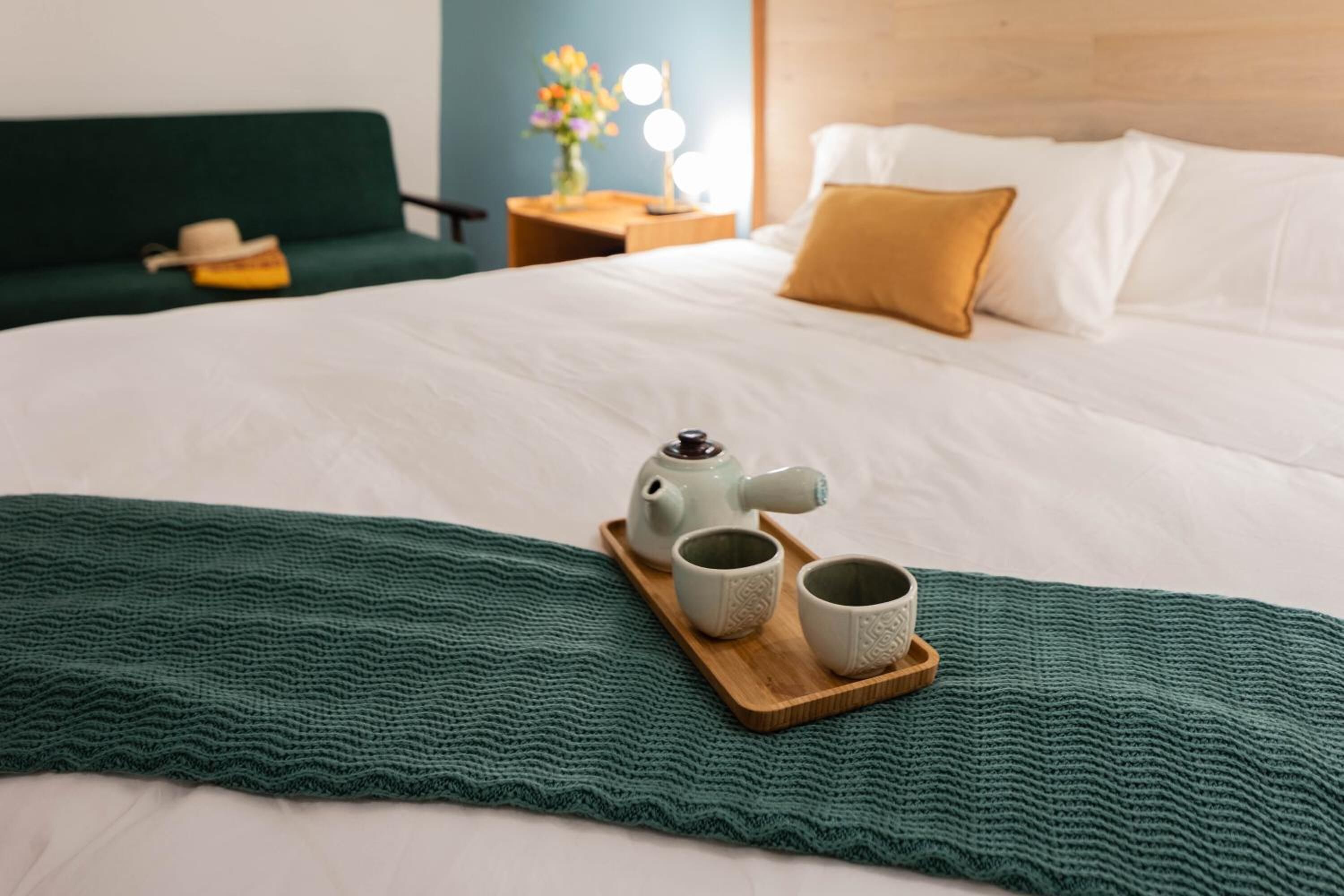 Una cama bien hecha con una manta verde, una bandeja de madera con un calentador y dos tazas, y un sofá verde al fondo.