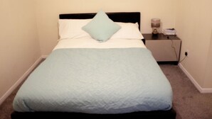 Un lit bien fait, avec une couverture et un oreiller bleu clair, une table de chevet avec une lampe, et une prise électrique sur le mur.