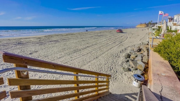 Een strand met een houten trap die naar het zand leidt, een rotsachtige kustlijn en een rode auto op het strand geparkeerd.