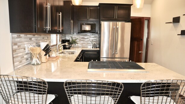 A modern kitchen with dark cabinets, stainless steel appliances, a marble countertop, and wire mesh chairs.