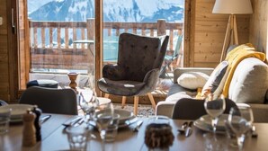 Un interior de madera con una mesa de comedor para dos personas, un sofá y una silla, con vistas a la montaña a través de grandes ventanales.