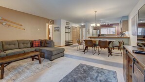 A spacious living room with a sectional sofa, a dining area with a long table and chairs, and a kitchen with a large window.