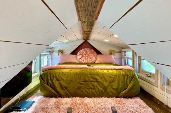 A cozy attic bedroom with a green bed, pink and white pillows, a fluffy rug, and a wooden ceiling.