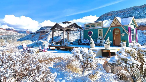 A snow-covered house with a porch, a small chapel, and a mountain backdrop. - Mother Eve Tiny House (Washington County)