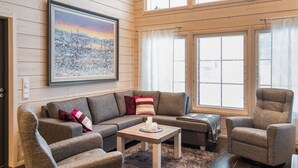A cozy living room with a sectional sofa, a small table, and a chair. There is a large window with blinds, a door, and a framed picture on the wall.