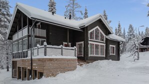 A large log cabin with a balcony and a covered porch, surrounded by snow-covered trees.