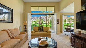 Un salon avec un canapé beige, une table basse en verre sur laquelle sont posés des livres, un téléviseur et une vue sur un terrain de golf, visible à travers de grandes fenêtres.