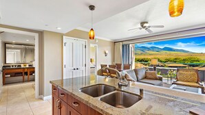 Una cocina moderna con encimeras de granito, fregaderos de acero inoxidable y armarios de madera. Hay una zona de comedor con un sofá y sillas, y un gran ventanal con vistas al paisaje.