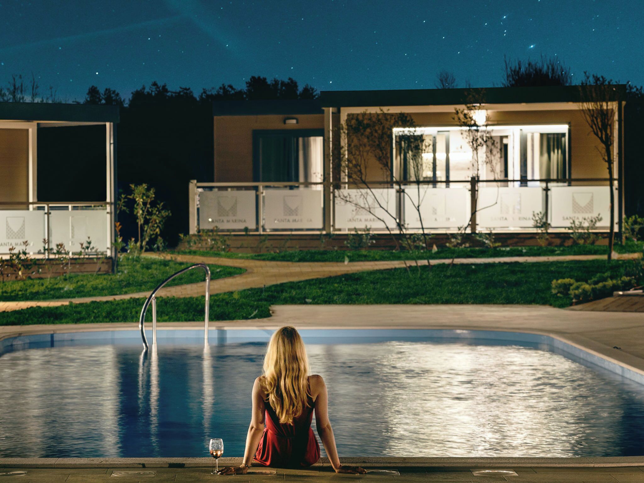 Eine Frau sitzt am Pool bei Nacht, mit einem Gebäude im Hintergrund.