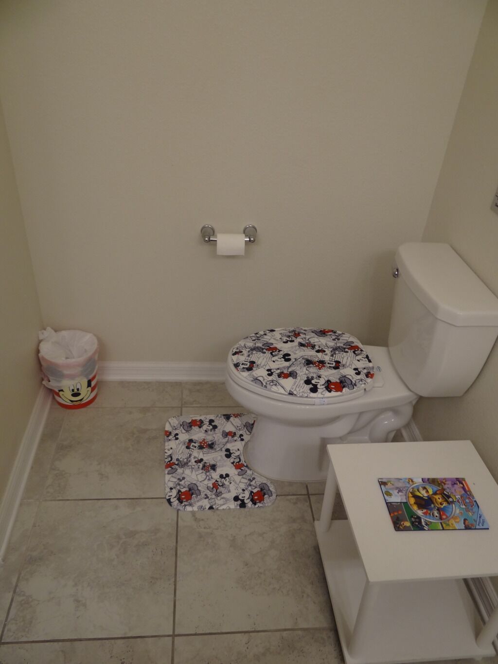 A small bathroom with a toilet, a roll of toilet paper, a Mickey Mouse trash can, and a small table with a children's book.