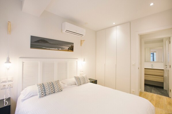 A bedroom with a large bed, white walls, a mounted air conditioner, a painting, and a wooden floor. - E1 APARTMENT WITH BALCONY IN SAN MARCIAL 33 (San Sebastián)