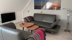 A modern living room with a grey leather sofa, a flat-screen TV, two wooden side tables, and a large framed picture of birds flying over a sunset. - Bird Migration - (Vollerwiek)