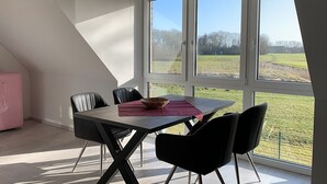 A dining area with a table and chairs, a pink cabinet, and a view of a green field through large windows. - Bird Migration - (Vollerwiek)