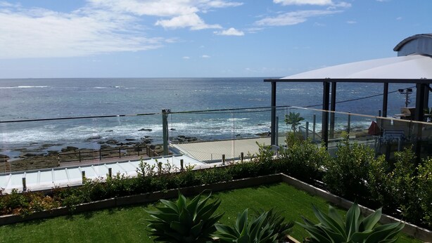 A coastal view from a balcony with a glass railing, a grassy area, and a white building with a curved roof. - Beachfront Penthouse The Entrance (The Entrance)