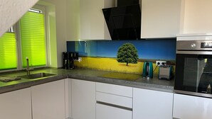 A modern kitchen with white cabinets, a built-in oven, and a backsplash with a landscape painting. - Dünentraum, Ferienwohnung in "De Feerjenhusen" am Haubarg von Vollerwiek (Vollerwiek)