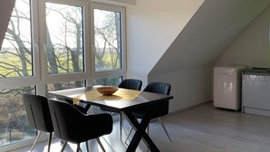 A dining area with a table and chairs, a small refrigerator, and a view of trees through large windows. - Dünentraum, Ferienwohnung in "De Feerjenhusen" am Haubarg von Vollerwiek (Vollerwiek)