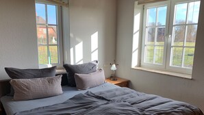 Une chambre à coucher avec un lit, des oreillers, une table de chevet, une lampe et une fenêtre donnant sur une maison et des arbres.