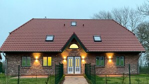 Un bâtiment en briques, surmonté d’un toit recouvert de tuiles rouges, avec deux fenêtres de part et d’autre, et une porte centrale dotée d’un petit auvent.
