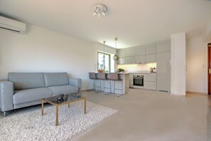 A modern living room with a grey sofa, a glass coffee table, and a white rug. There is a kitchen area with white cabinets and a window. - Modern Family Home 5 min to Beach +Pool +Garage.                                (Antibes)