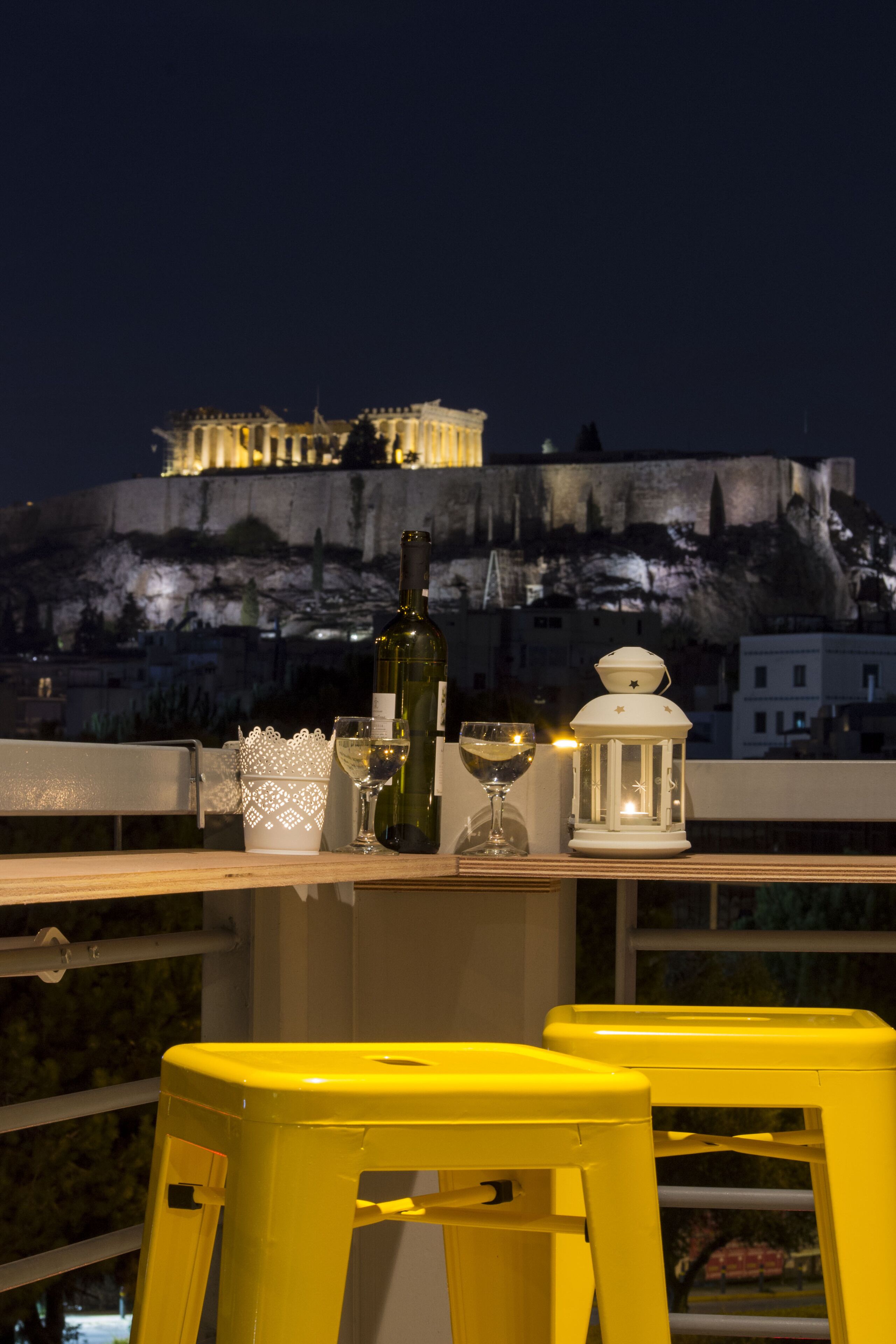 Uma mesa com uma garrafa de vinho, um copo e um lanterna branca numa varanda com vista para a cidade à noite.