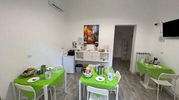 Ein Essbereich mit zwei Tischen, jeder Tisch mit einer grünen Tischdecke und weißen Stühlen. Auf den Tischen stehen Teller, Tassen und ein Korb. Darüber an der Wand hängt ein Gemälde.