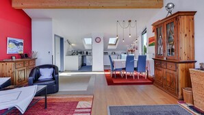 A modern living room with a red accent wall, a dining area with blue chairs, and a kitchen in the background.