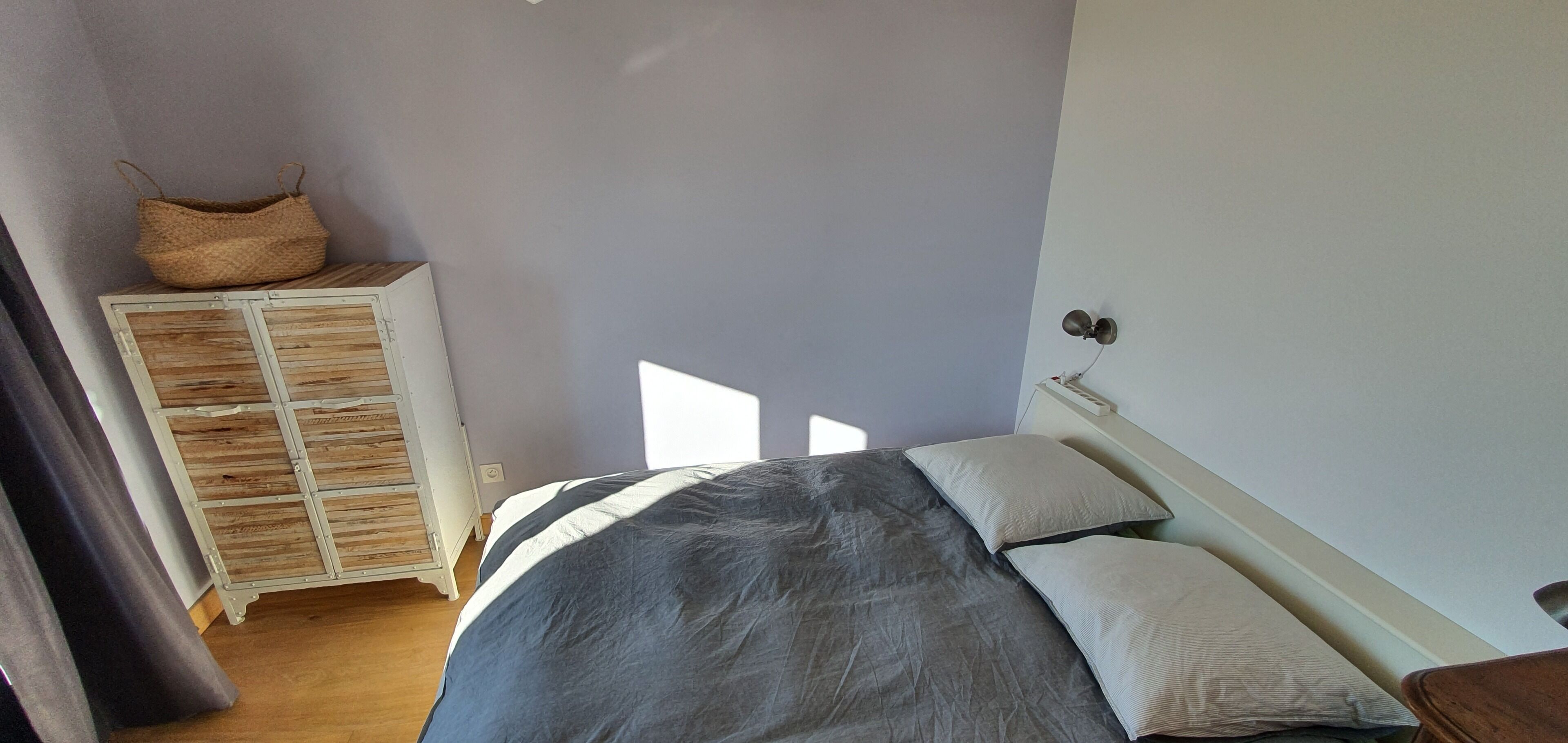 A bedroom with a bed, pillows, a nightstand, a lamp, a wardrobe, and a woven basket on top.