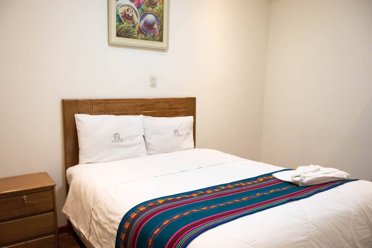 Una cama bien hecha con sábanas blancas y una manta a rayas de colores, una mesita de noche de madera con cajón, y un cuadro en la pared.