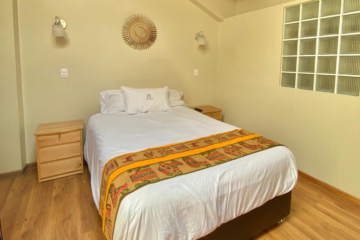 Un dormitorio con techo de madera, una cama con colcha blanca, una mesita de noche, una lámpara de pared y una ventana con persianas.