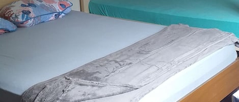 A single bed with a grey blanket, a lower bunk bed with a floral mattress, and a shelf with a red cup and a green bottle.