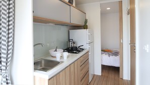 Une cuisine compacte équipée d’un évier, d’une cuisinière et de meubles blancs. On aperçoit un lit recouvert d’une couvre-lit à motifs floraux dans la pièce adjacente.